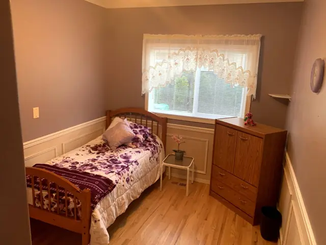 A private resident bedroom with a twin bed, warm hardwood floors, a dresser, and a lace-curtained window